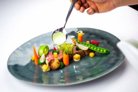 Koch von Markus Grein Catering gibt bei einem Event grüne Soße auf ein Gericht aus Fleisch, Erbsen und Gemüse.