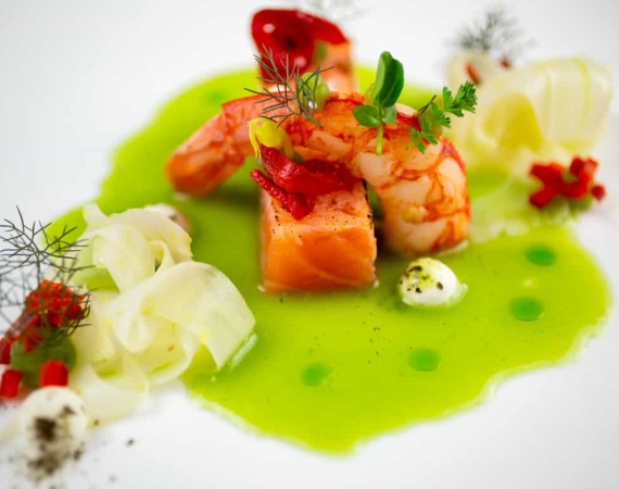 Fein angerichteter Lachs mit Garnele, Gemüsestreifen und grüner Soße auf einem weißen Teller.