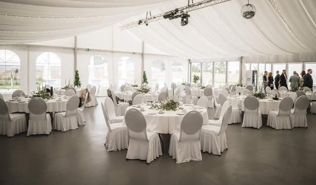 Mehrere runde Tische mit Hochzeitsdekoration und Stühle mit Hussen in einem Pavillon der Eventlocation 2Event in Wiesentheid.