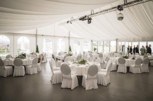 Mehrere runde Tische mit Hochzeitsdekoration und Stühle mit Hussen in einem Pavillon der Eventlocation 2Event in Wiesentheid.
