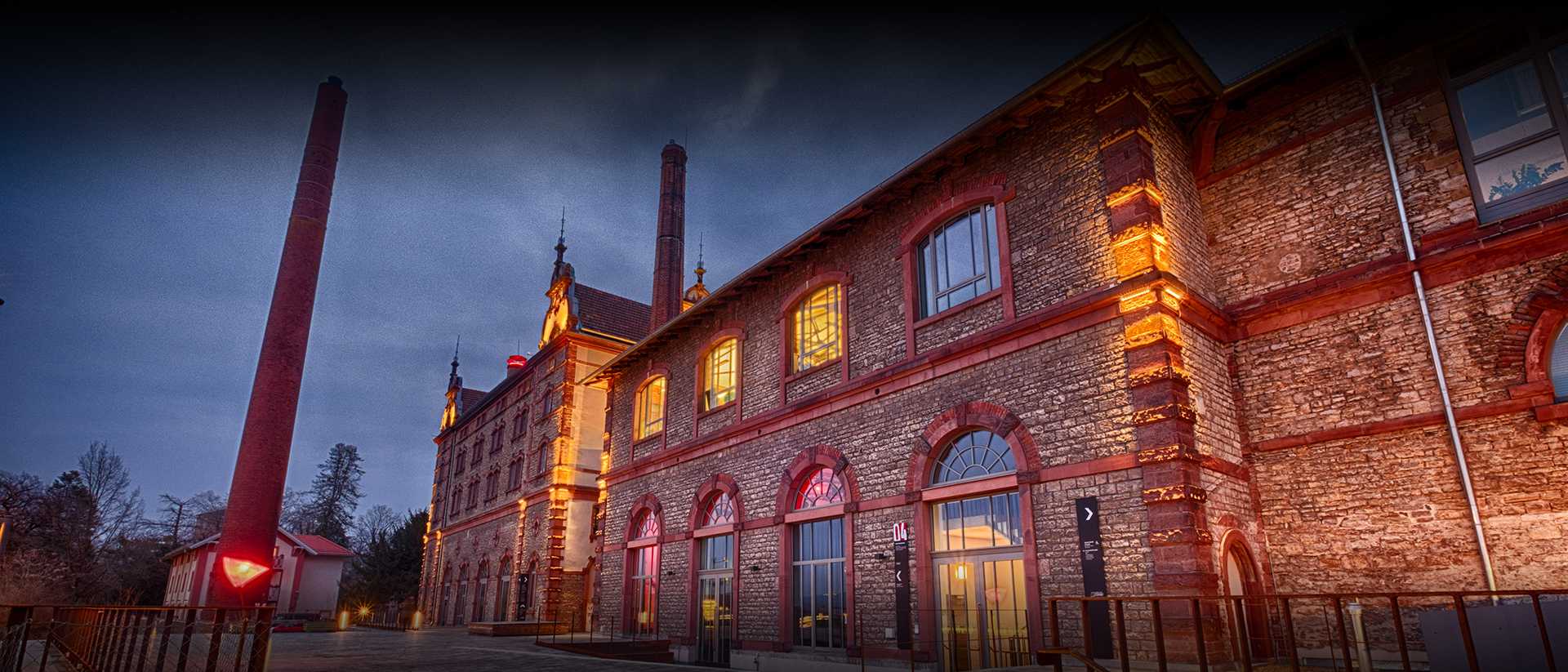 Beleuchteter Außenbereich der Würzburger Eventlocation im Industriestil, Maschinenhaus der Bürgerbräu bei Abenddämmerung.