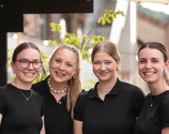 Vier junge Mitarbeiterinnen im Service lächeln gemeinsam in die Kamera bei einem Event im Außenbereich