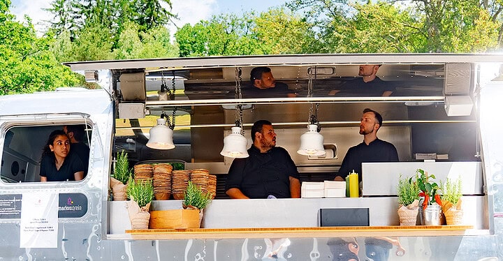 Team beim Foodtruck Catering hinter der geöffneten Klappe eines silbernen Foodtrucks im Grünen