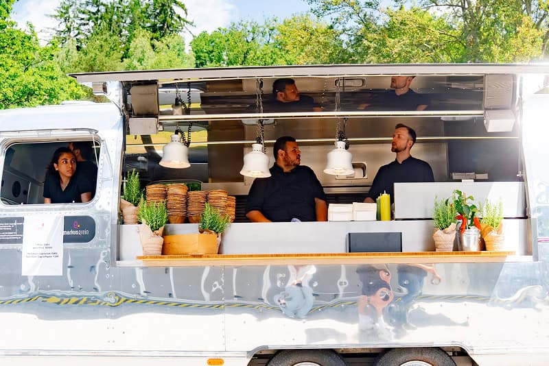 Team beim Foodtruck Catering hinter der geöffneten Klappe eines silbernen Foodtrucks im Grünen