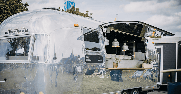 Silberner Airstream-Foodtruck steht für das Messe-Catering auf einer grünen Wiese bereit.