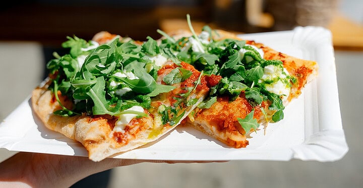 Hand hält einen Pappteller mit einem Stück Pizza mit frischem Rucola beim Pizza Catering auf einer Hochzeit