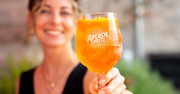 Servicekraft von Markus Grein Catering aus Würzburg zeigt bei einem DJ-Event von vamos. ein Glas Aperol Spritz in die Kamera
